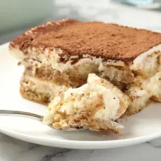 Kawałek najprostszego tiramisu na talerzu, z widocznymi warstwami biszkoptów i kremu. Na łyżeczce porcja deseru.