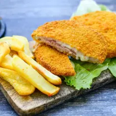 Chrupiący kotlet po szwajcarsku z rozpływającym się serem i szynką, podany z frytkami na desce.