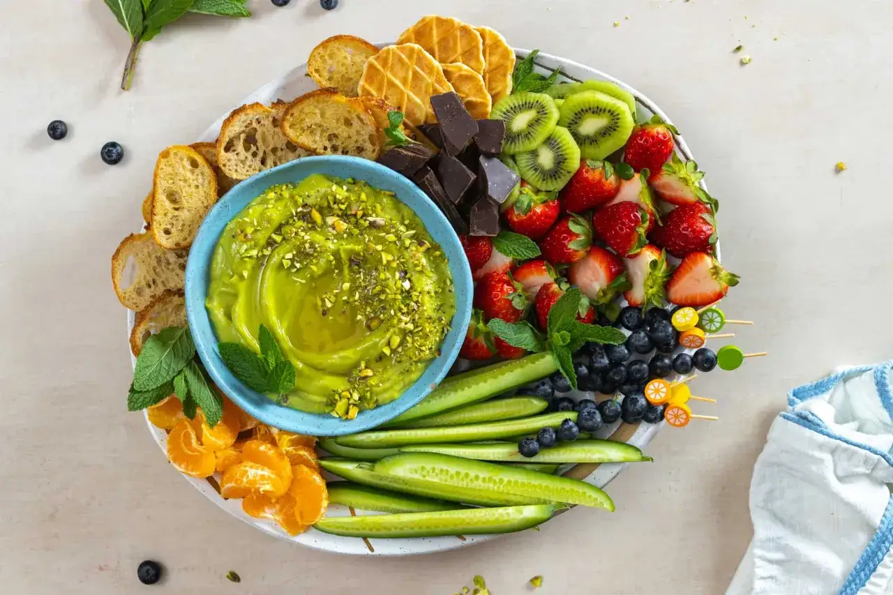 Kolorowa deska z pysznym, zielonym dipem, który wygląda jak guacamole. Obok świeże owoce, warzywa i pieczywo. Idealny przepis na guacamole!
